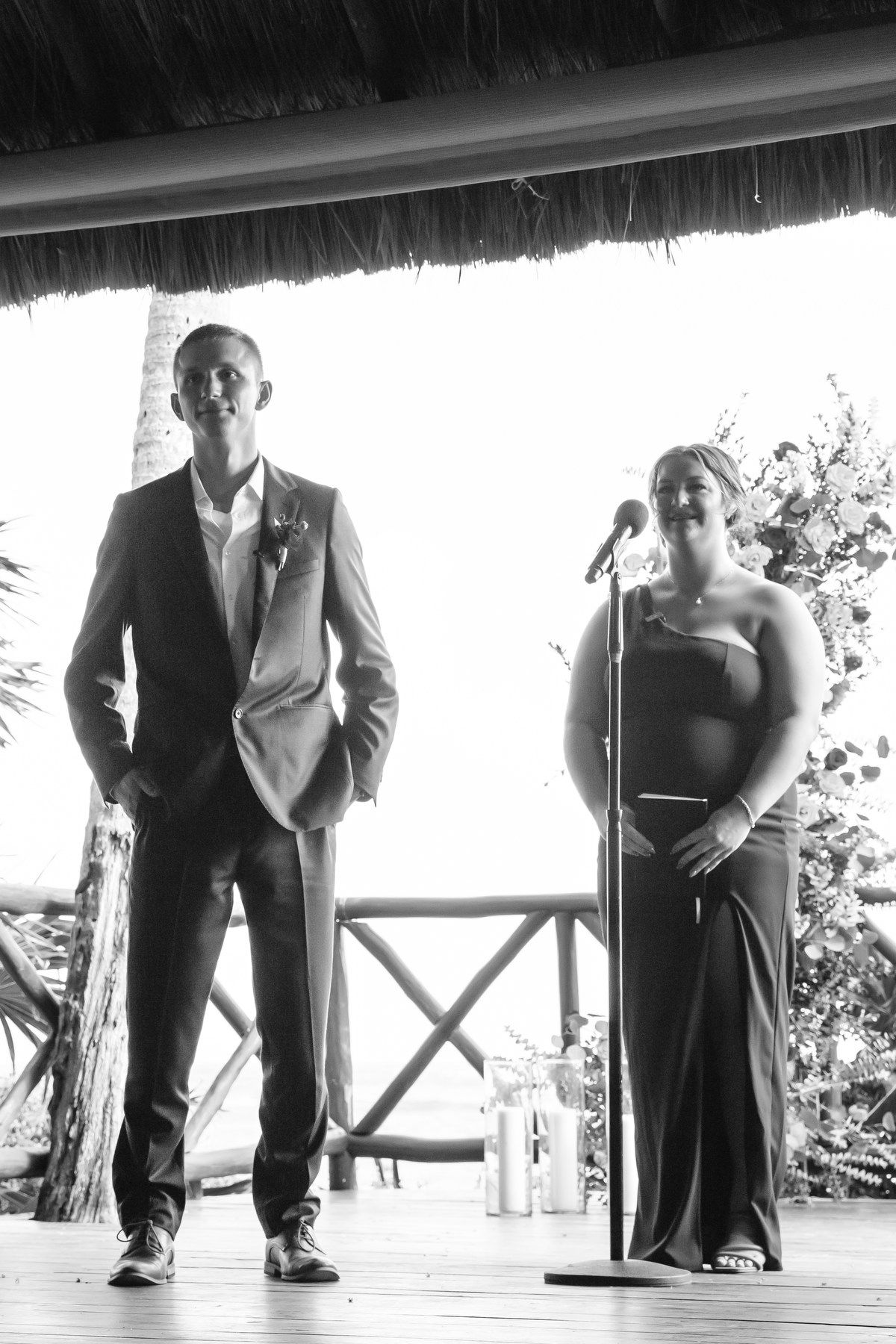 Groom stands at the altar as a bridesmaid speaks at a microphone during an outdoor beach wedding ceremony.