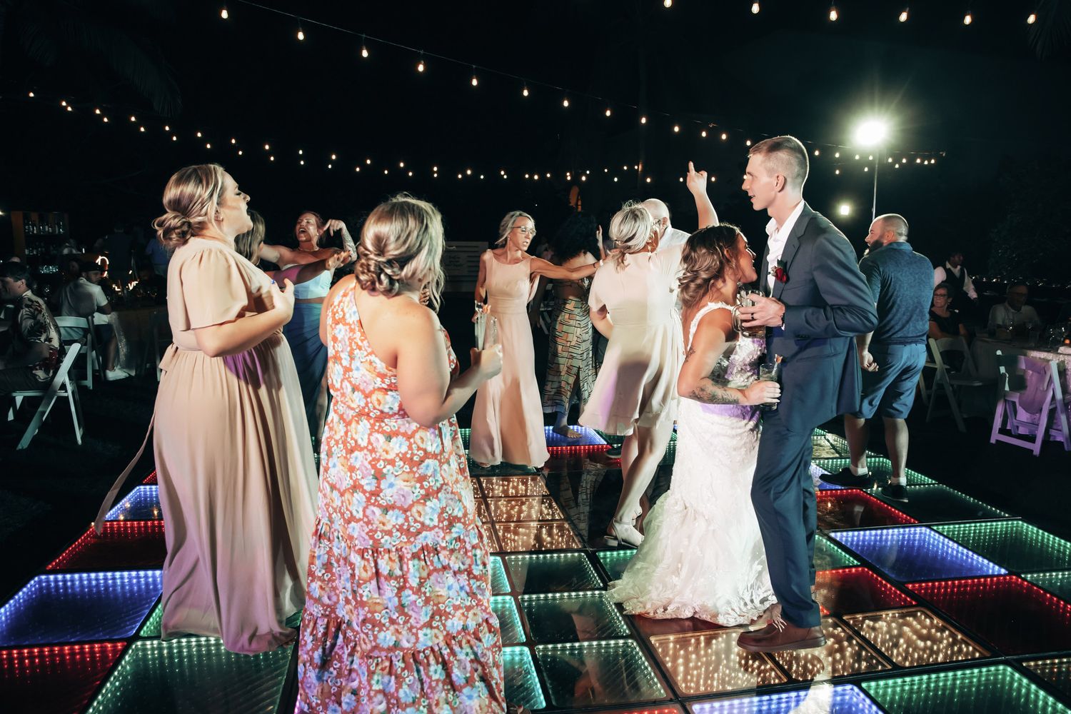 Wedding guests and bridal party dancing on colorful LED dance floor under string lights at night reception.
