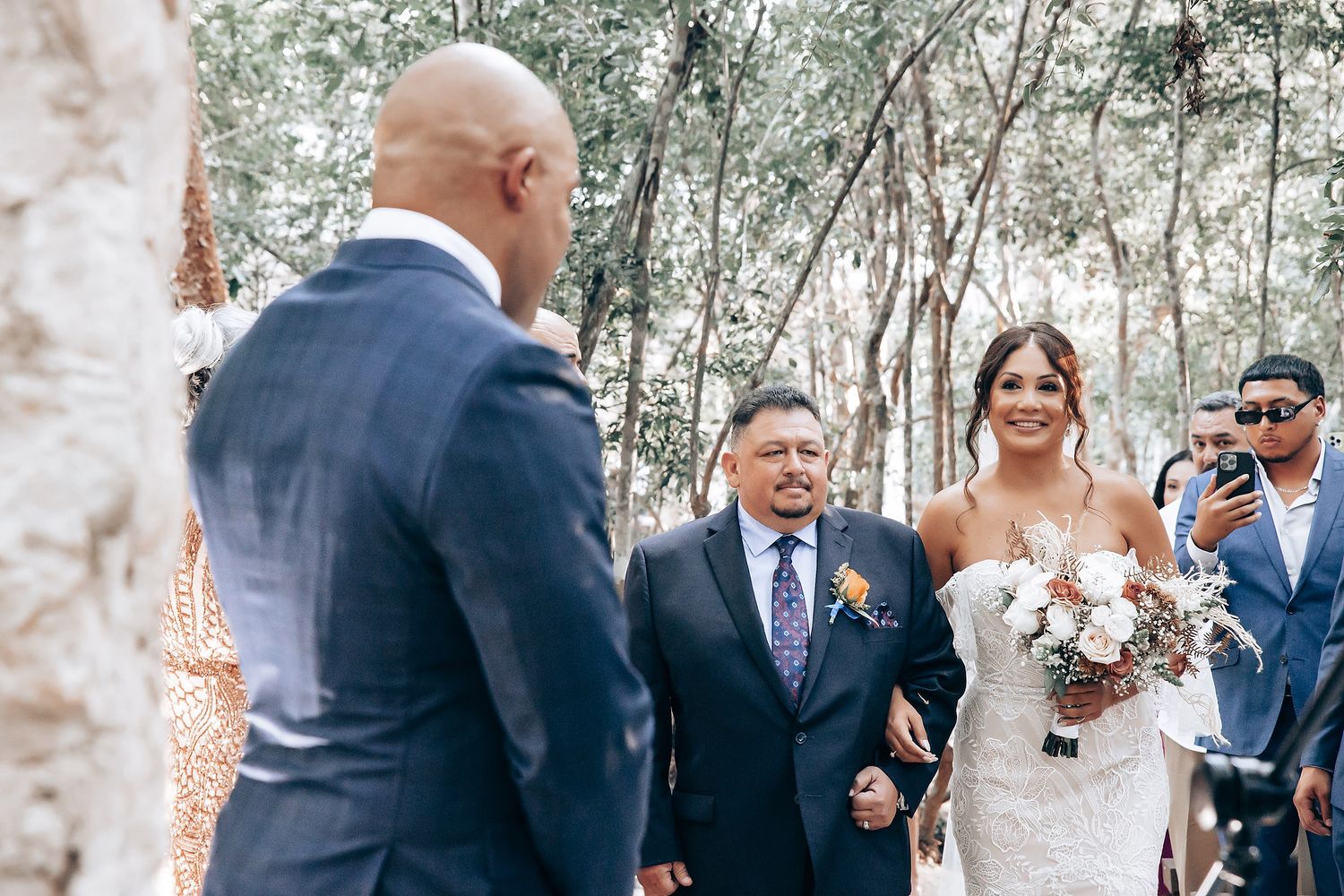 Wedding ceremony guests gathered under trees in an outdoor setting.