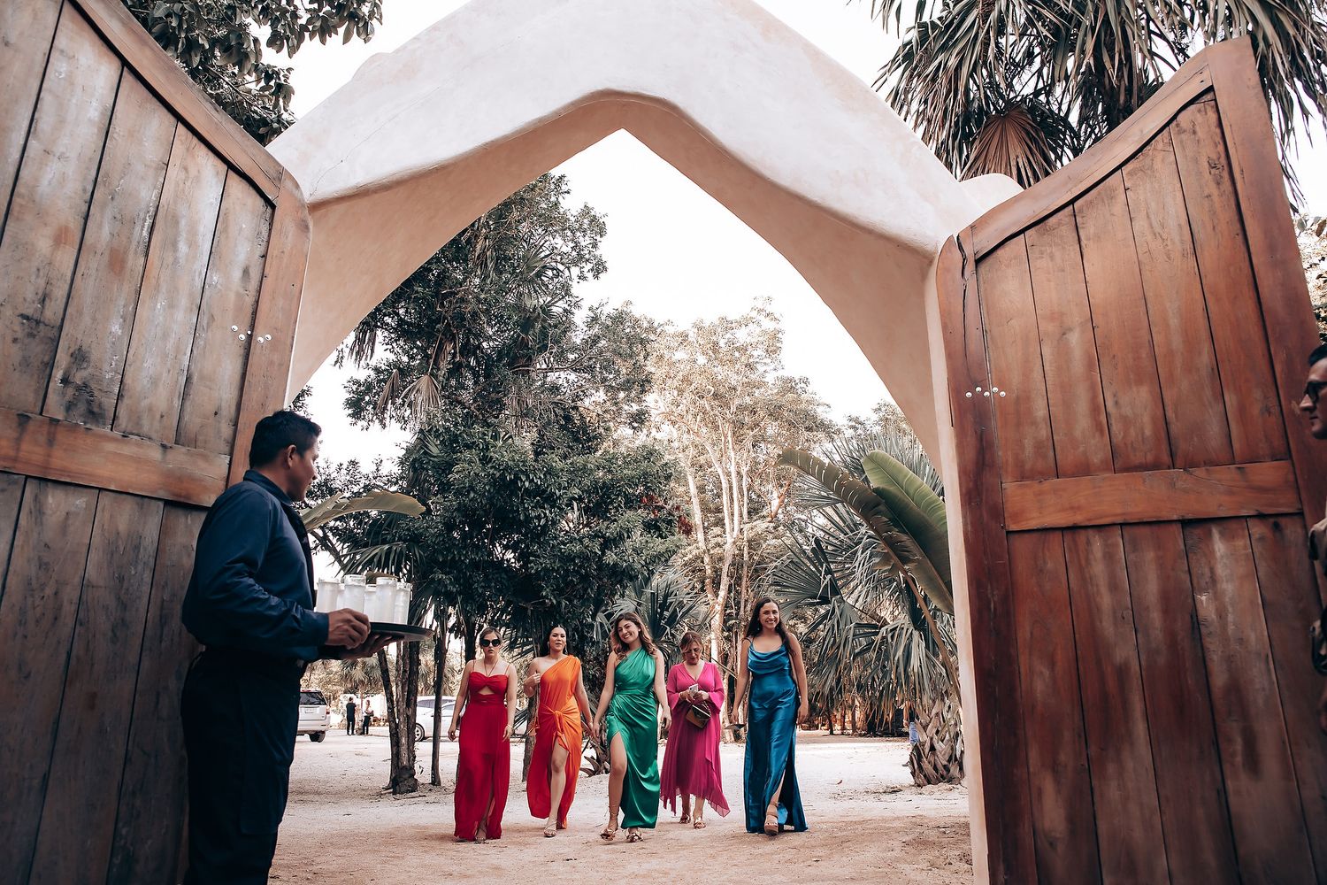 Wedding party walks along a rustic wooden path surrounded by tall trees at an outdoor venue.