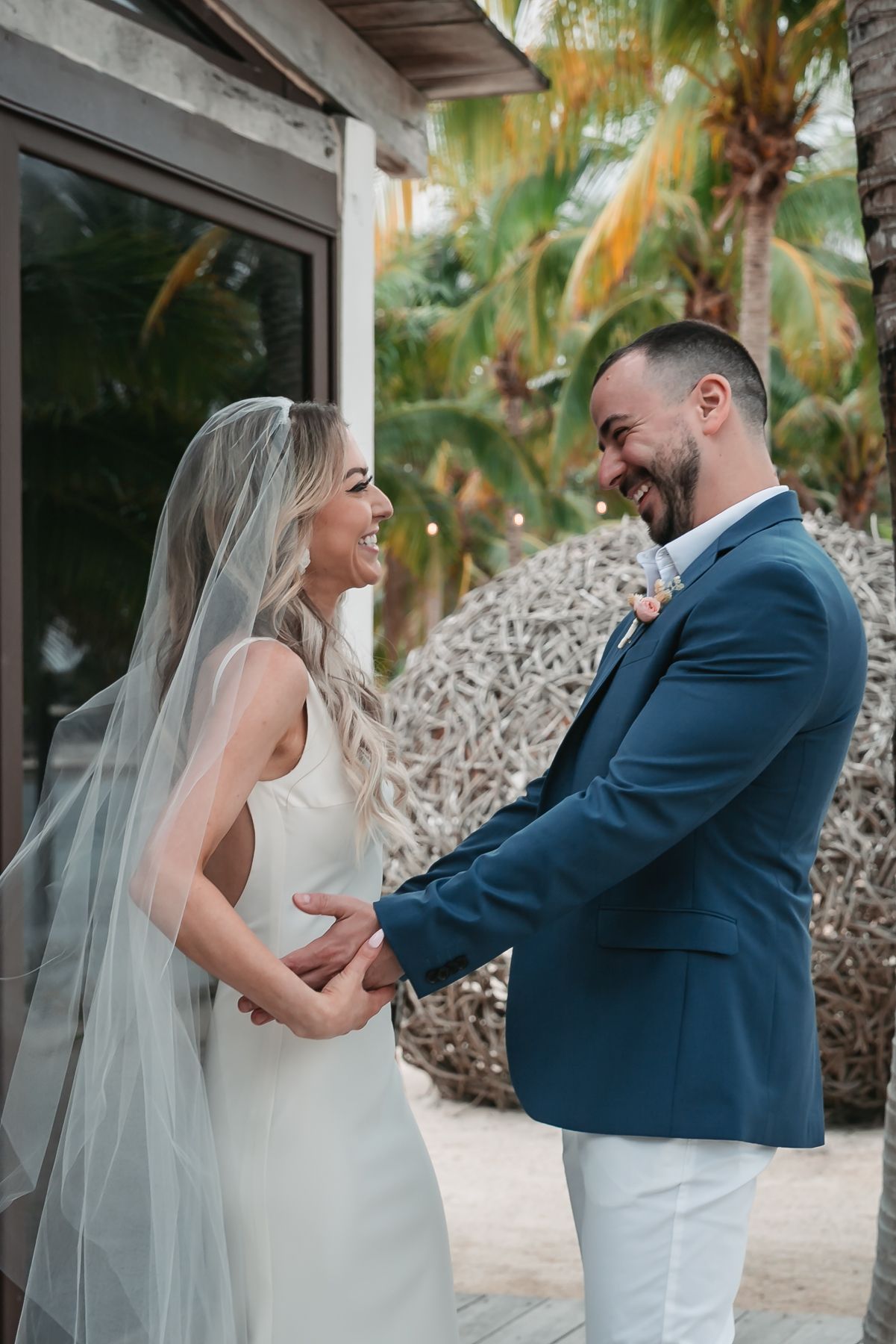 Joyful bride and groom holding hands and laughing together outdoors at a tropical beach wedding venue.