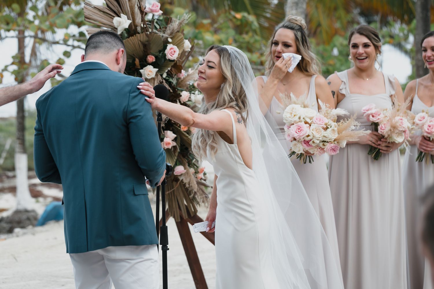 Emotional bride touching grooms face at beach wedding ceremony with bridesmaids in taupe gowns.