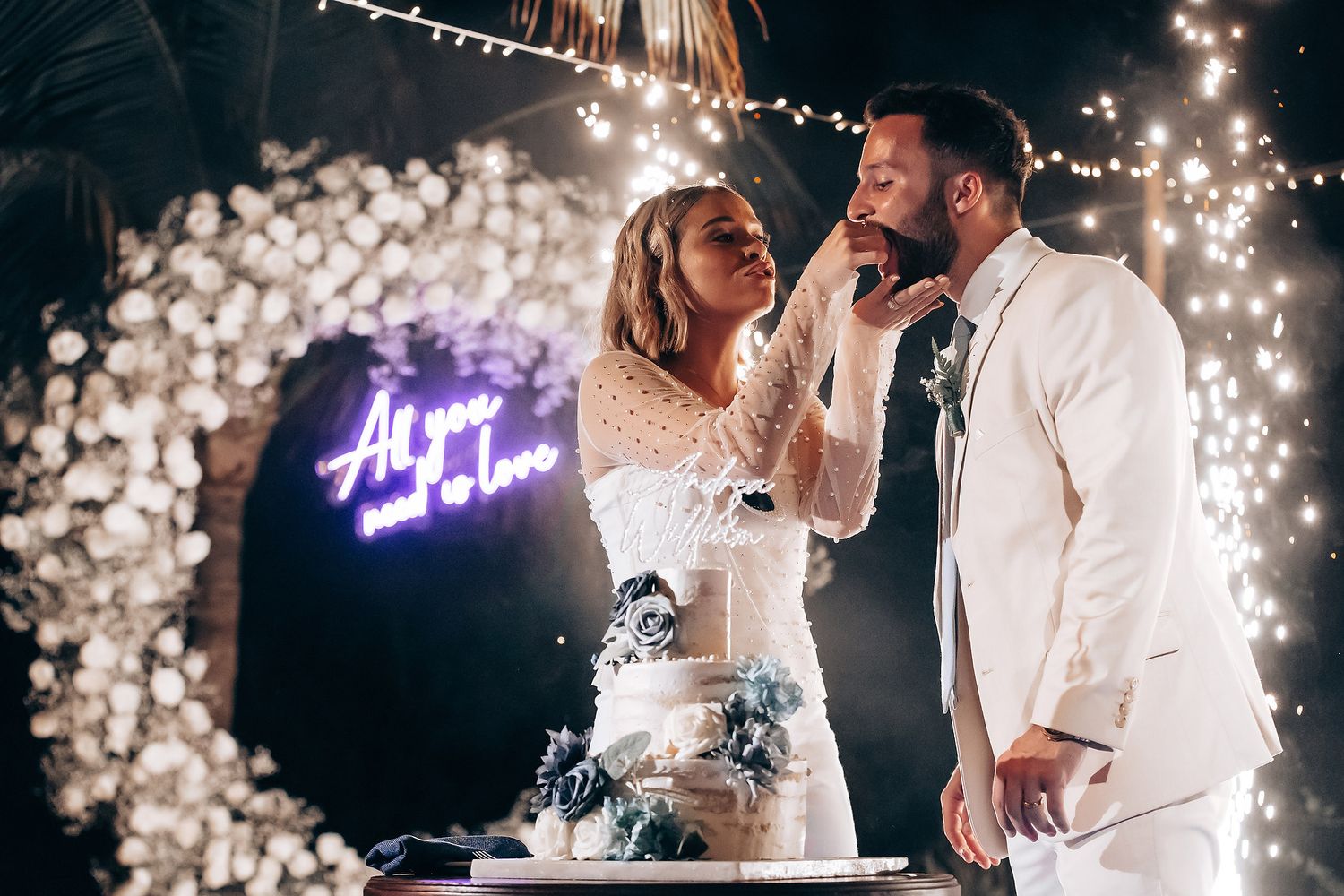 A couple shares a romantic moment while cutting their wedding cake beneath twinkling lights and a neon sign.