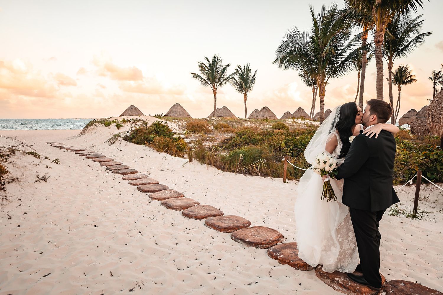 Wedding couple walks along a tropical beach pathway at sunset.