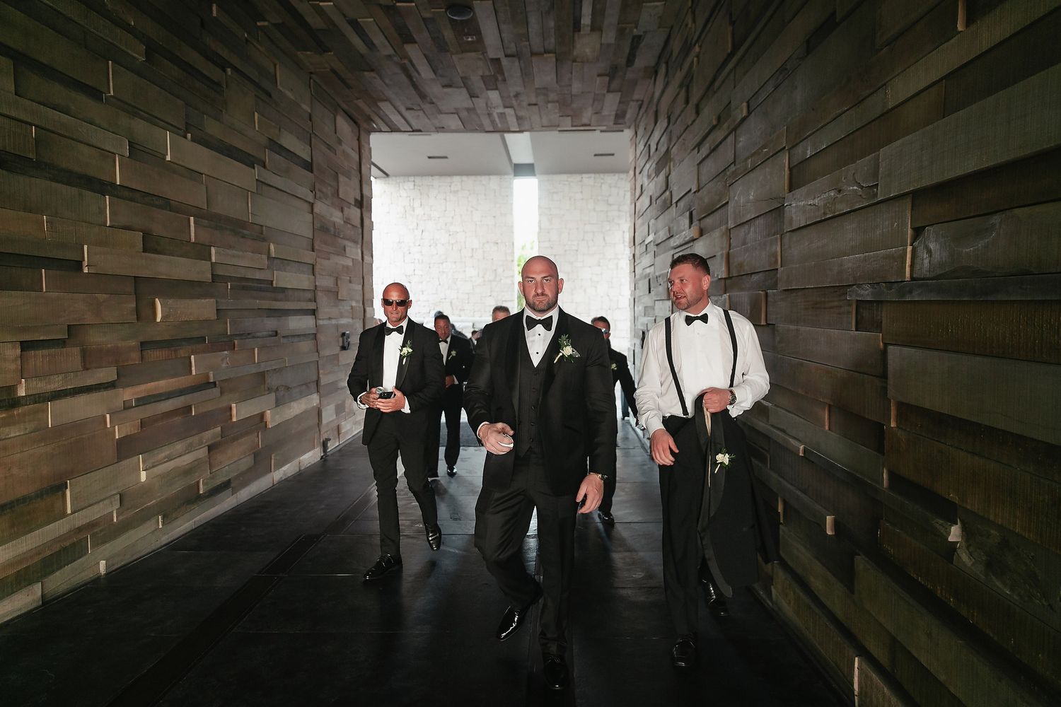 Groomsmen in tuxedos and suspenders pose in a dramatic dark wooden hallway for wedding photos.