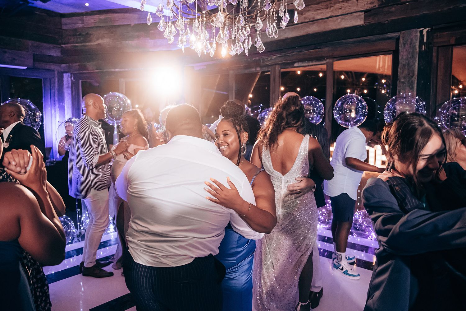 Wedding guests dance and celebrate on a dimly lit dance floor under elegant crystal chandeliers during a reception.