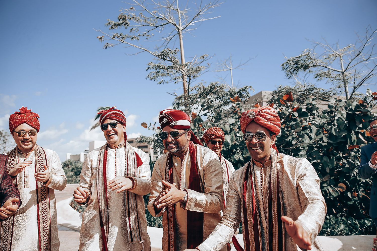 Groomsmen in matching cream sherwanis and red turbans celebrating outdoors.