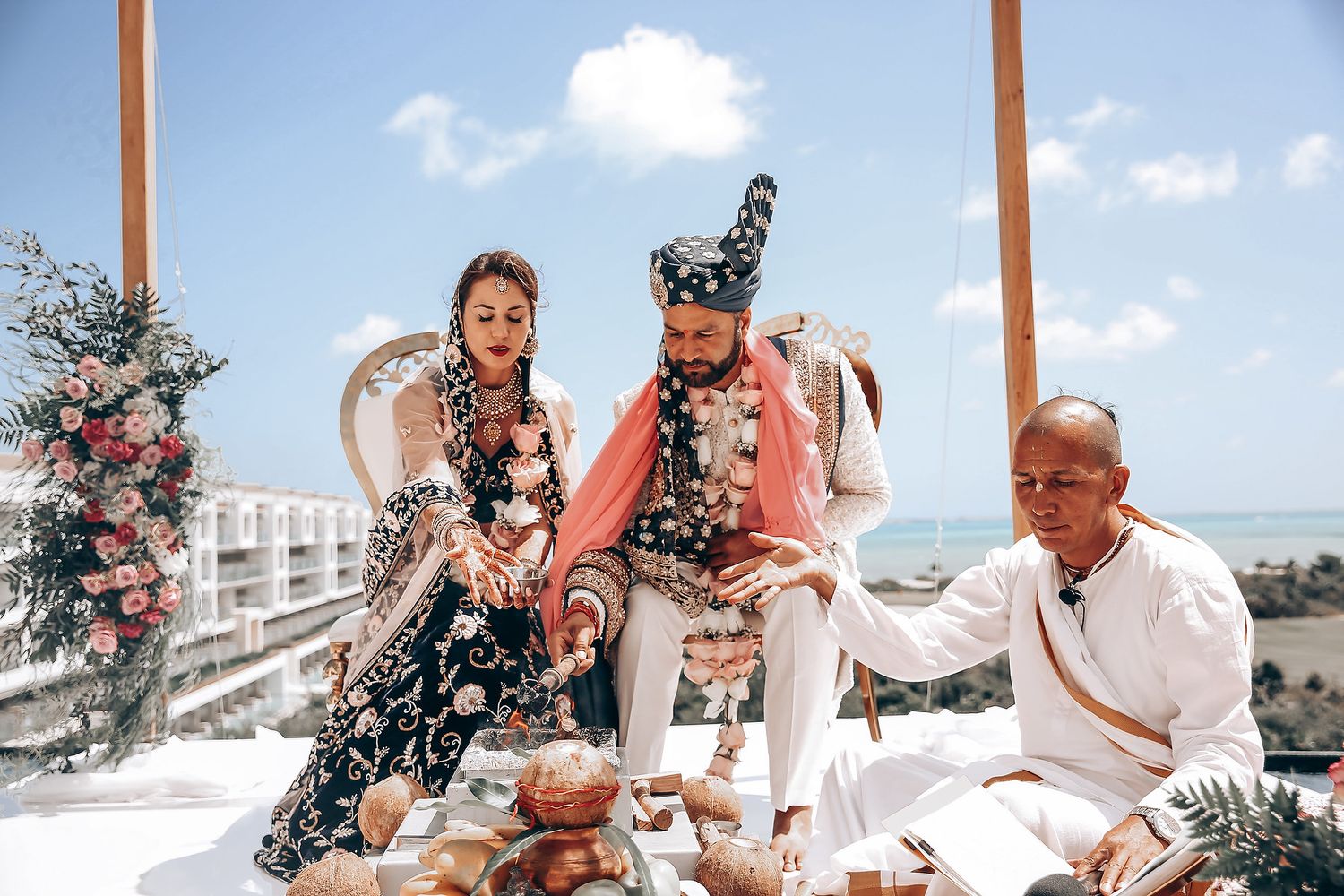 Guests gather for a ceremonial moment at a beachside wedding under wooden posts decorated with pink flowers.