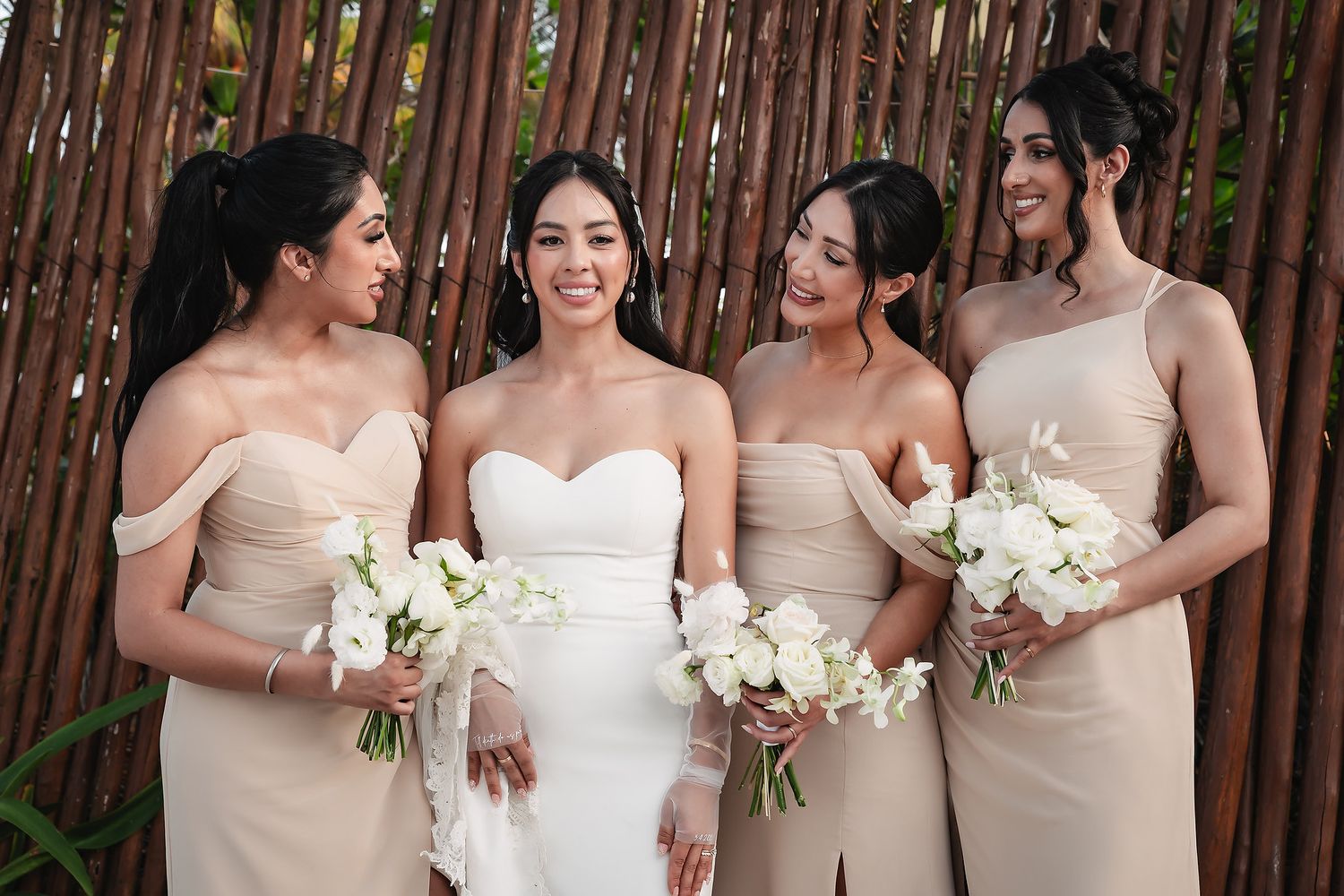 Wedding party in neutral tones holding white bouquets against bamboo wall backdrop.