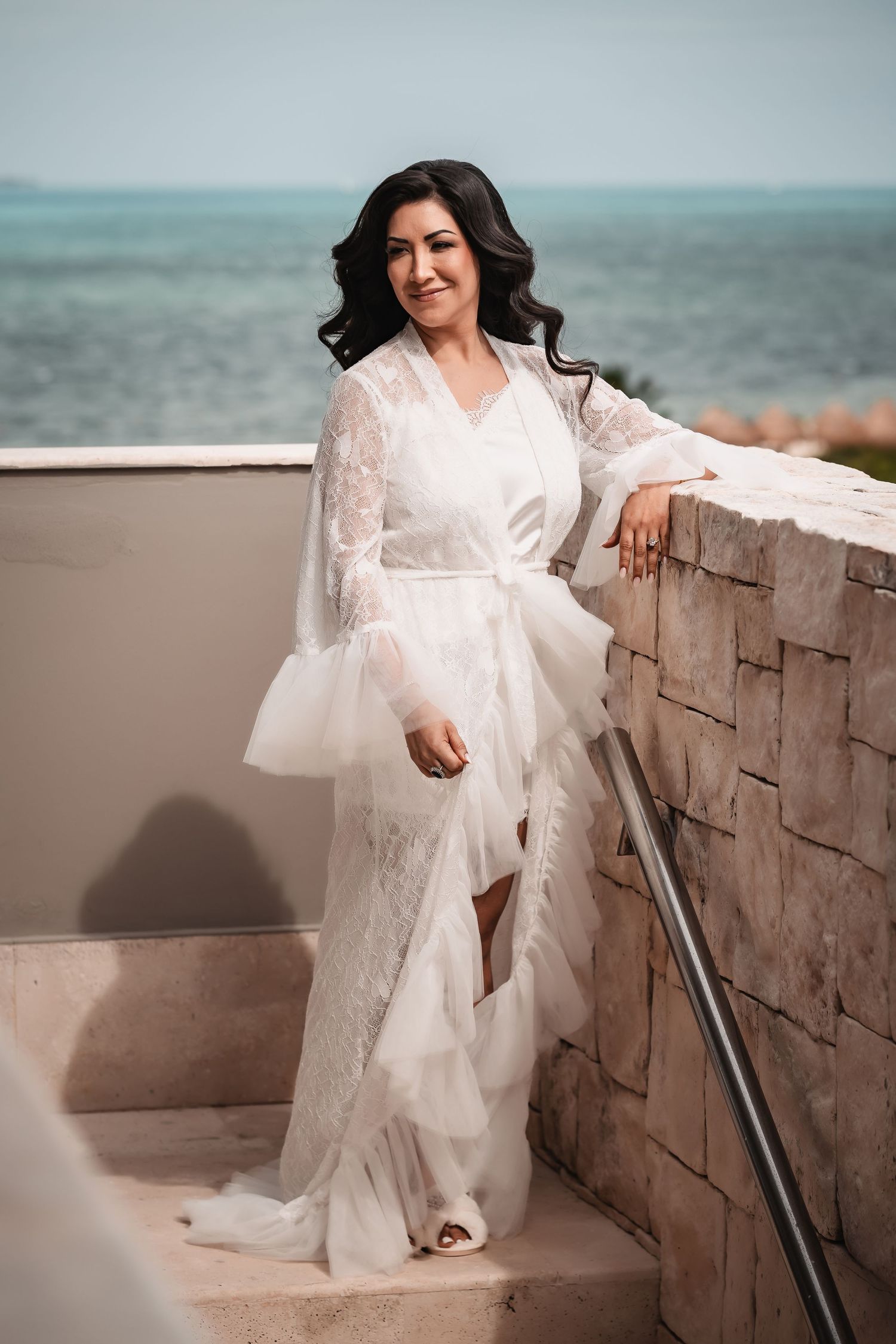A white lace wedding dress flows elegantly as someone poses on stone steps near the ocean.
