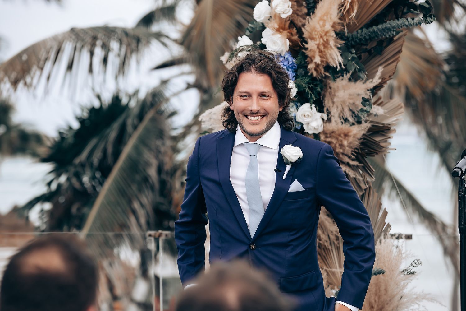 Wedding ceremony guests seated outdoors with palm trees and floral decorations in a tropical setting.