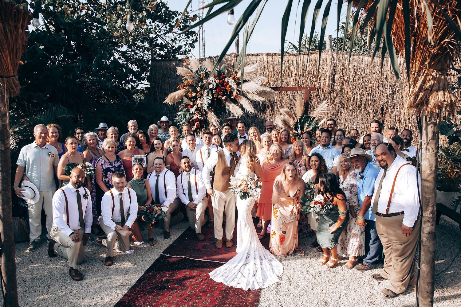 A series of wedding guests line up for a panoramic group photo outdoors at a rustic venue.