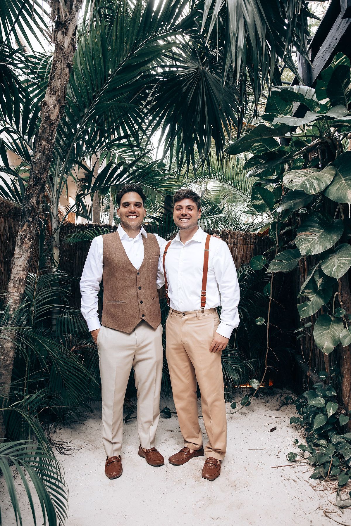 Two people in coordinated vintage-style outfits with suspenders stand together in a lush tropical garden setting.
