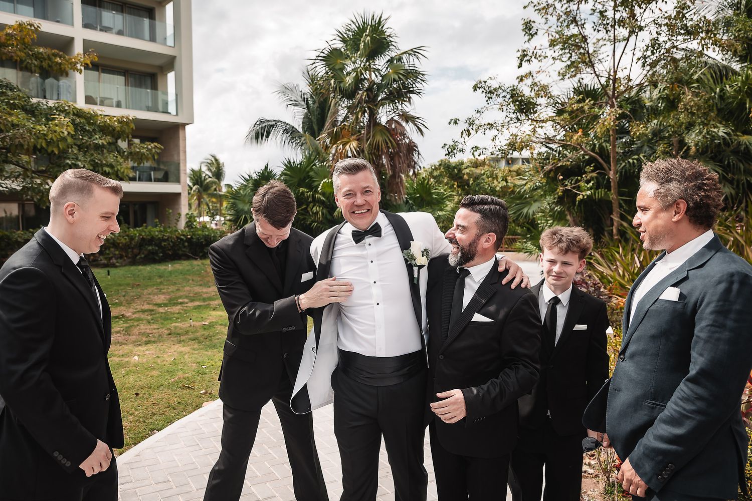 Groomsmen in black tuxedos laughing and celebrating together outdoors.