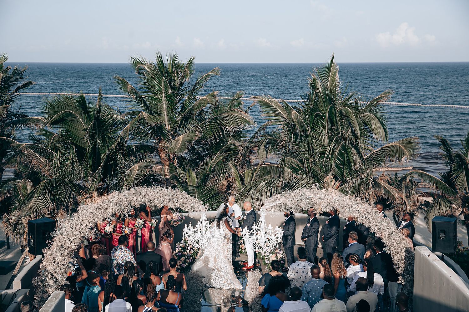 Oceanfront wedding ceremony under a floral arch with palm trees and seated guests.