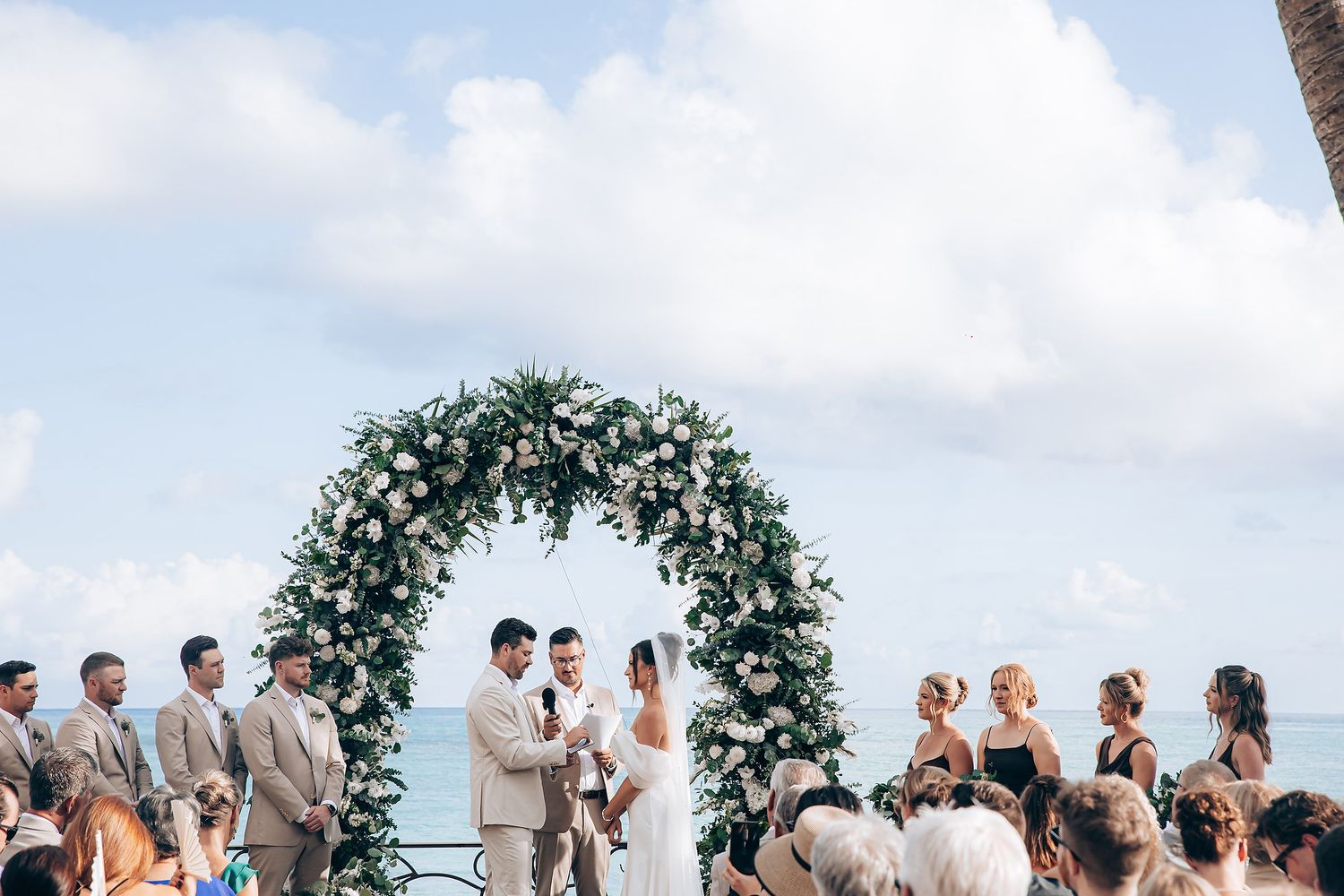 A wedding ceremony setup on a beach with guests gathered under blue skies.