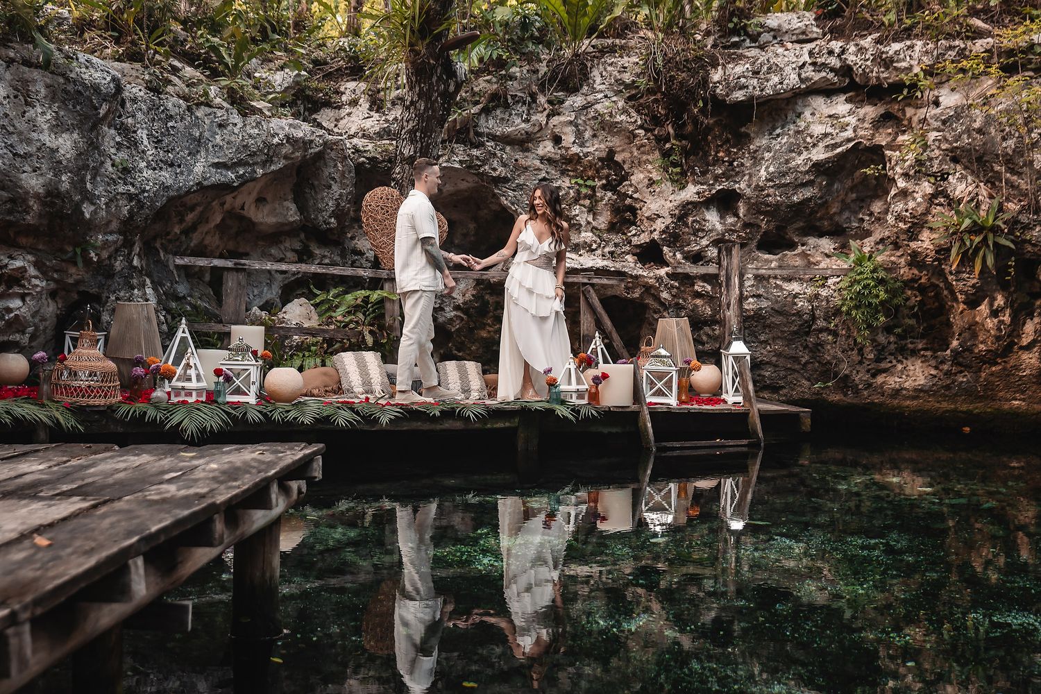 Couple standing on wooden dock by crystal clear cenote waters surrounded by limestone cliffs.