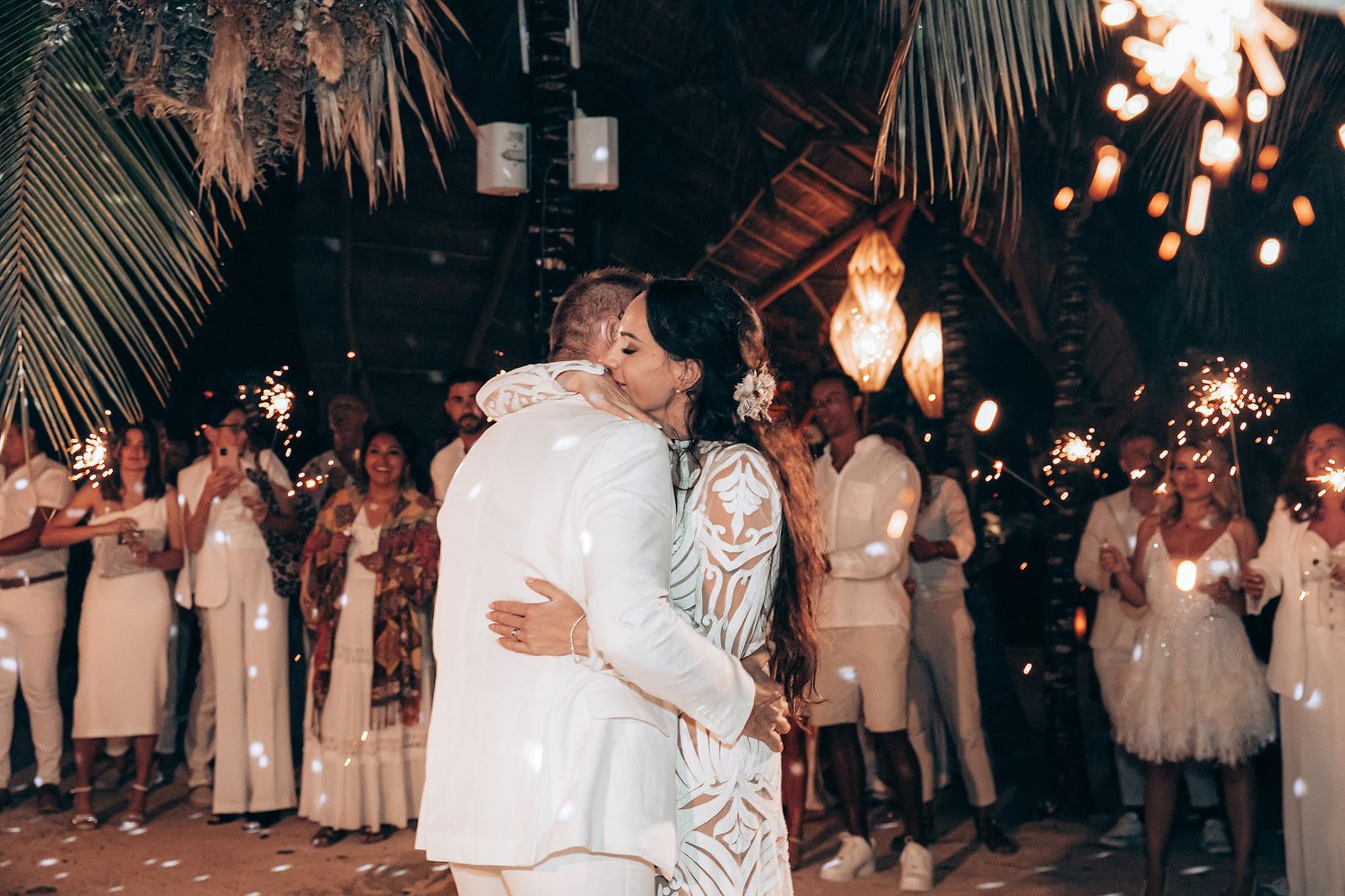 Wedding guests dance under palm trees and string lights at an outdoor evening reception celebration.