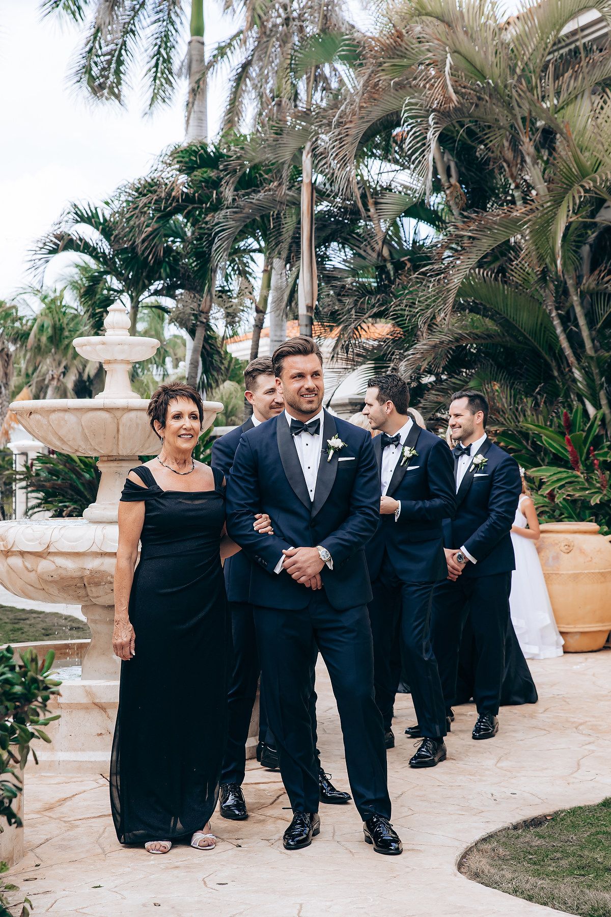 Wedding party members in black dresses and tuxedos share candid moments while posing on outdoor stone patio.