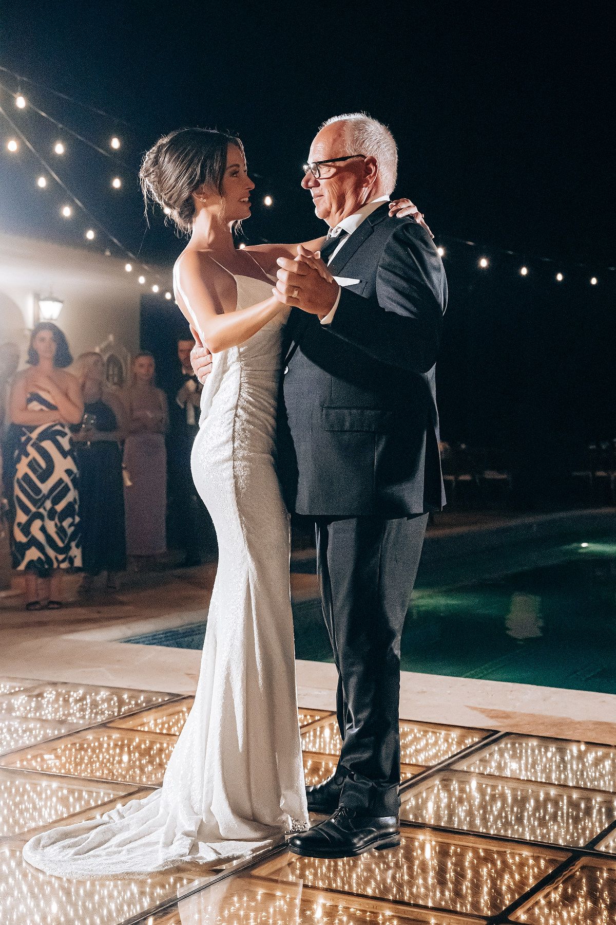 A couple shares a tender dance moment on an illuminated dance floor under string lights at an evening celebration.