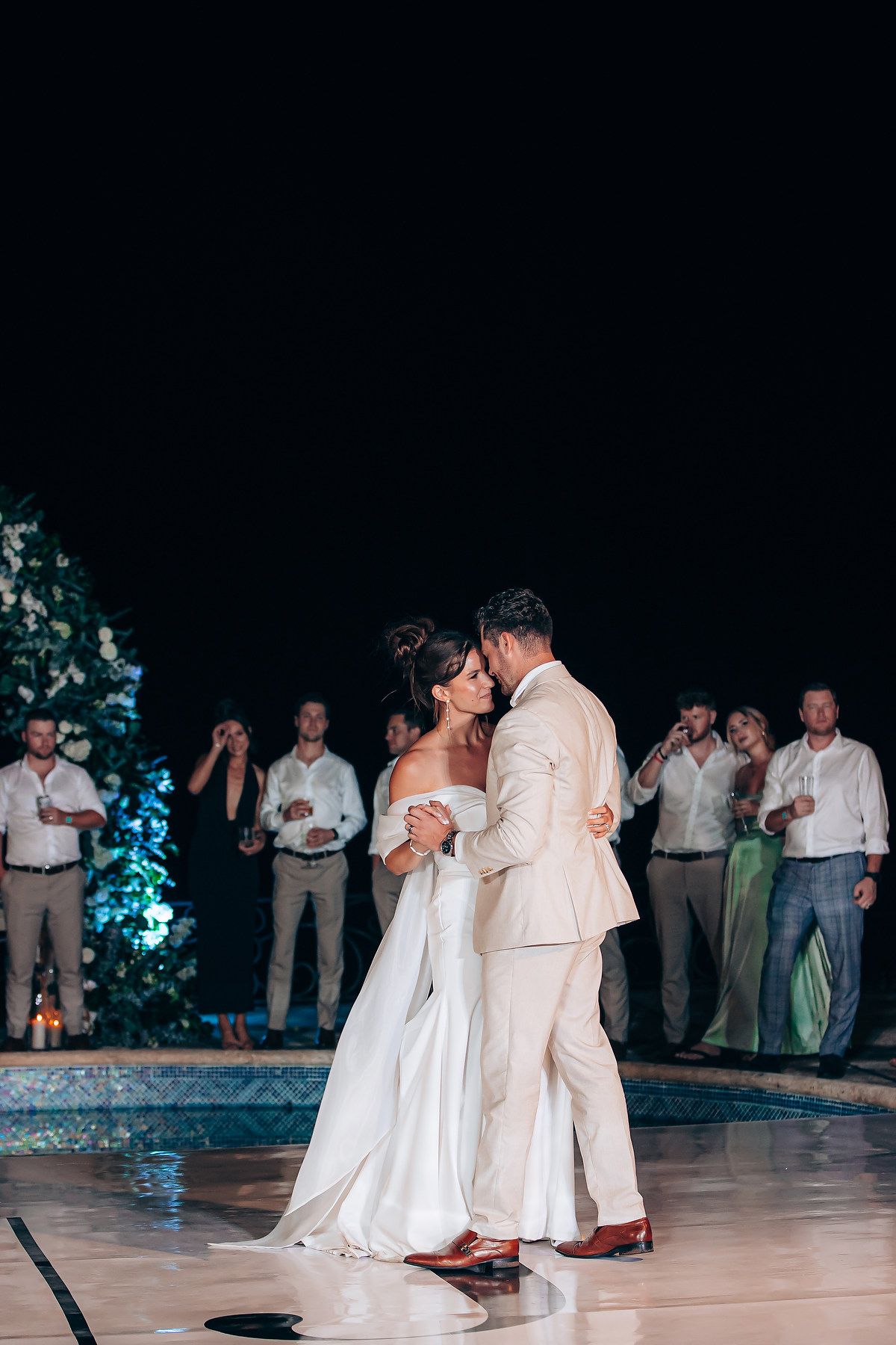 Couple shares their first dance as newlyweds on an outdoor dance floor at night with guests watching in the background.