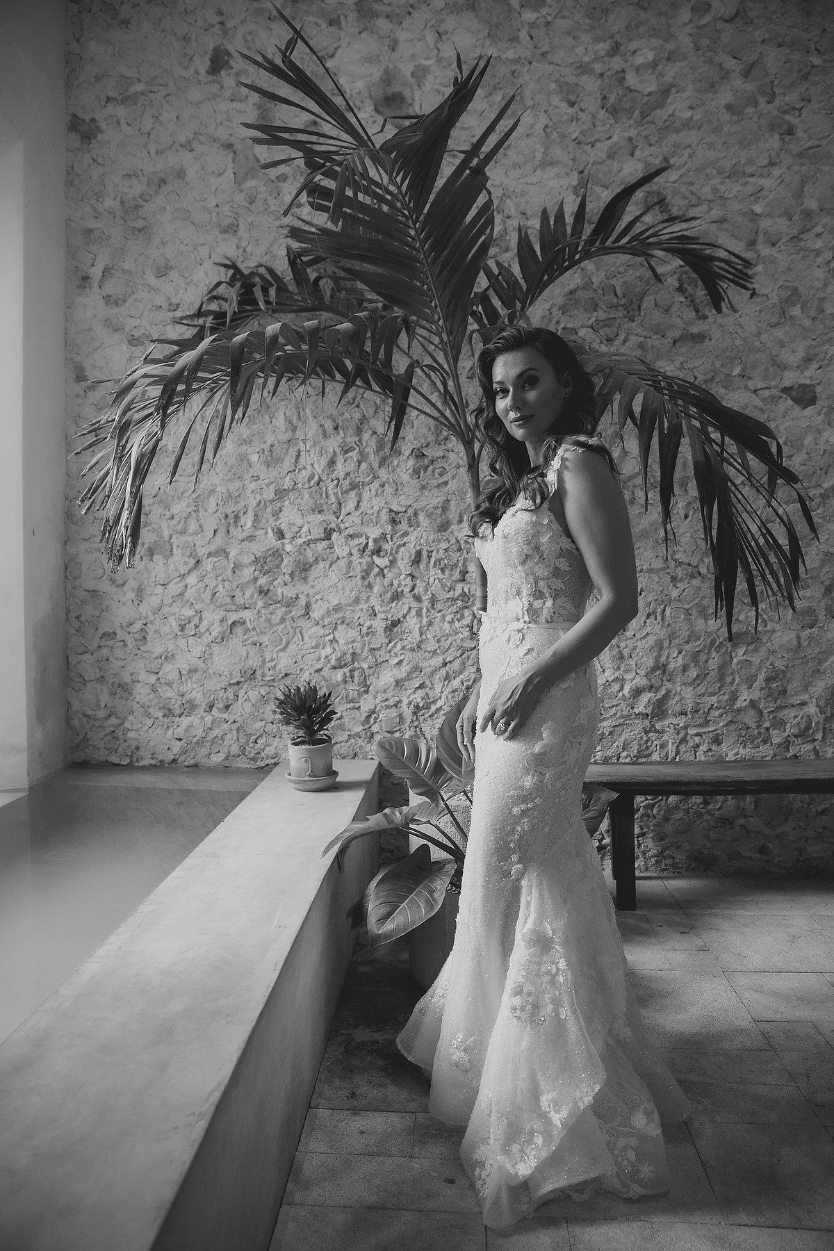 A bride in a white lace wedding dress twirls and dances in front of a rustic stone wall with tropical palm plants.