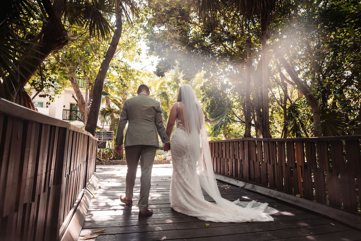 Natural light streams through trees onto a wooden walkway during a wedding ceremony.