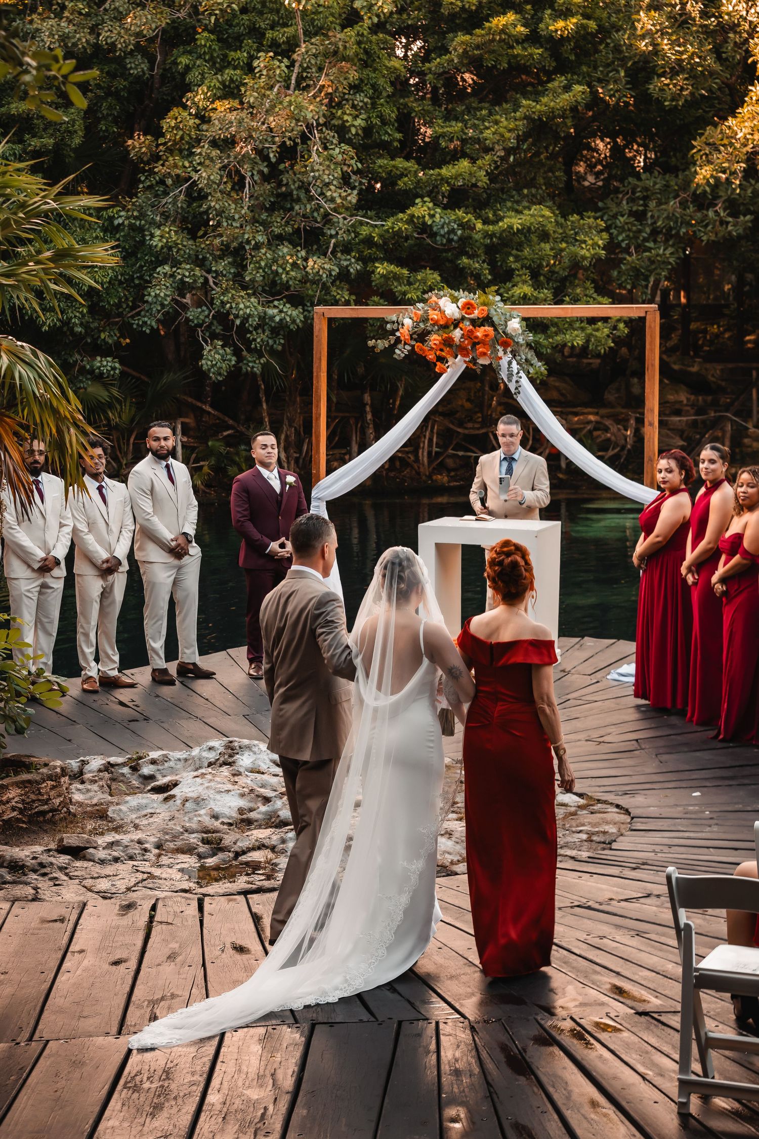 Outdoor wedding ceremony setup with wooden arch, white draping, and burgundy bridesmaids on wooden deck.