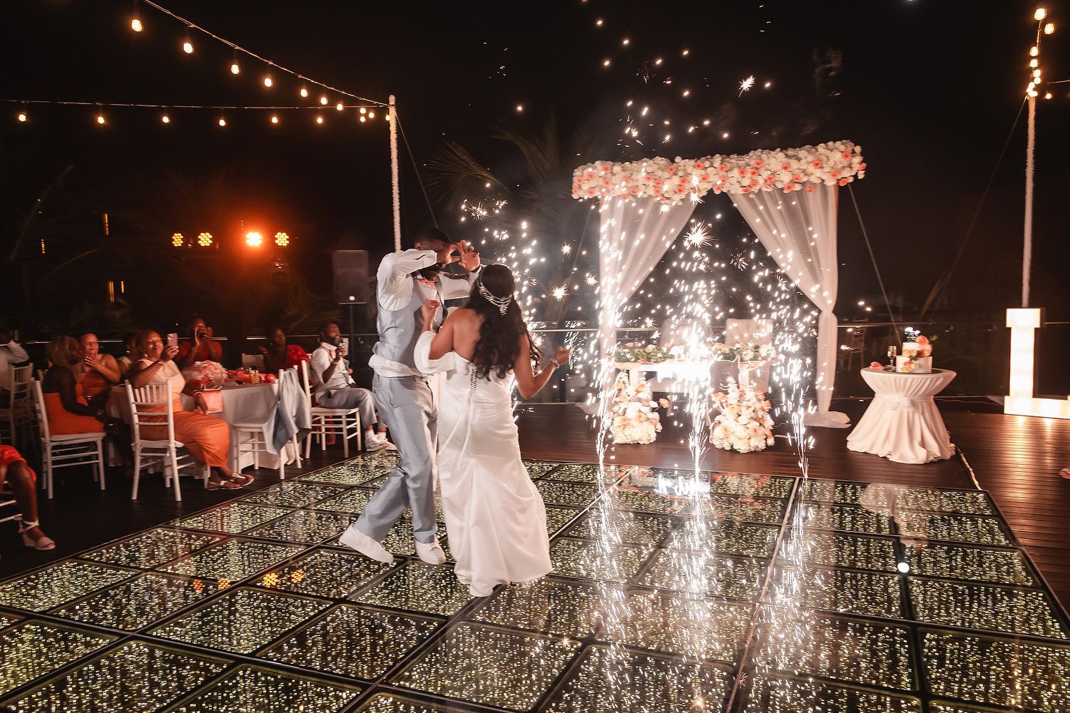 Couple's first dance under string lights with sparklers and floral arch at night.