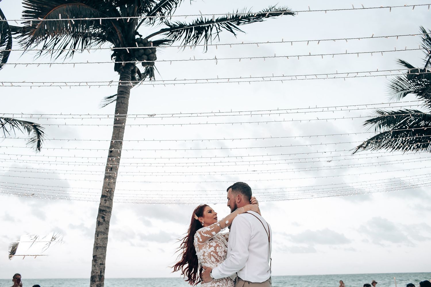A couple shares a romantic kiss on a tropical beach with palm trees and cloudy skies in the background.