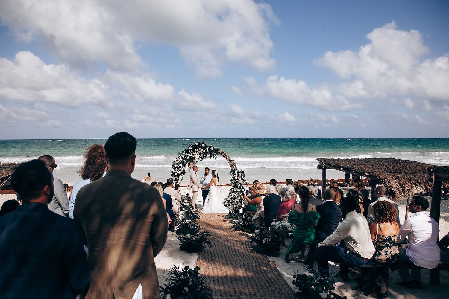Wide view of beach wedding ceremony with floral arch and seated guests on wooden pathway.