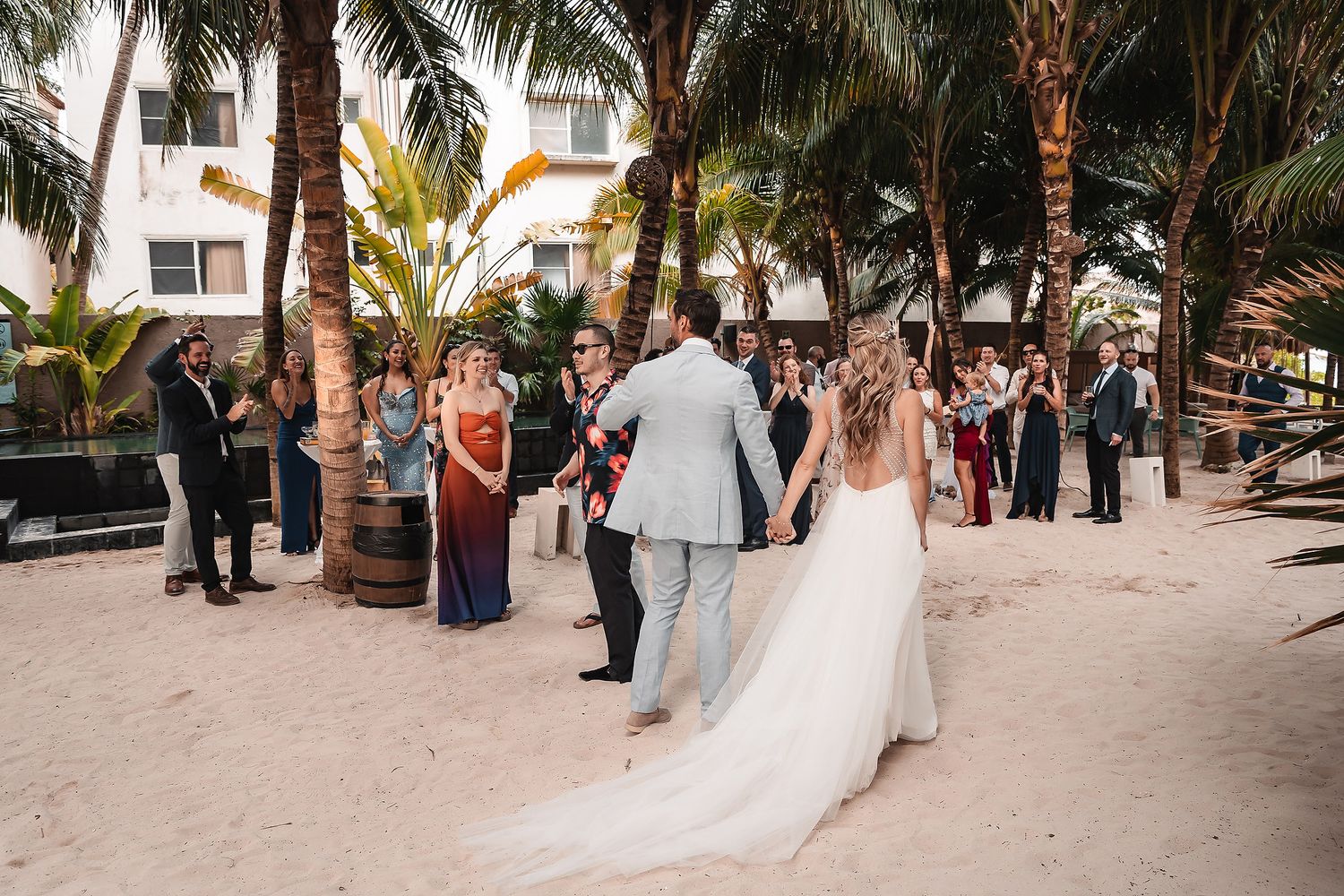 Wedding guests gather on a sandy beach path lined with palm trees during a destination ceremony.
