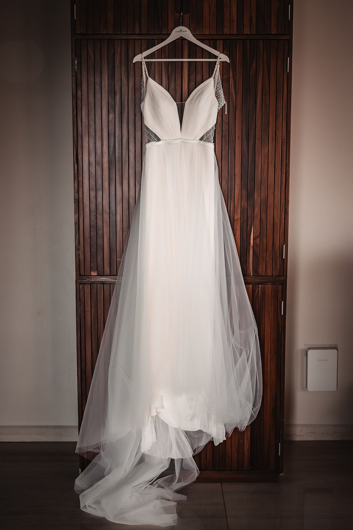 A white wedding dress hangs elegantly against a dark wooden backdrop, showcasing its flowing tulle skirt and plunging neckline.