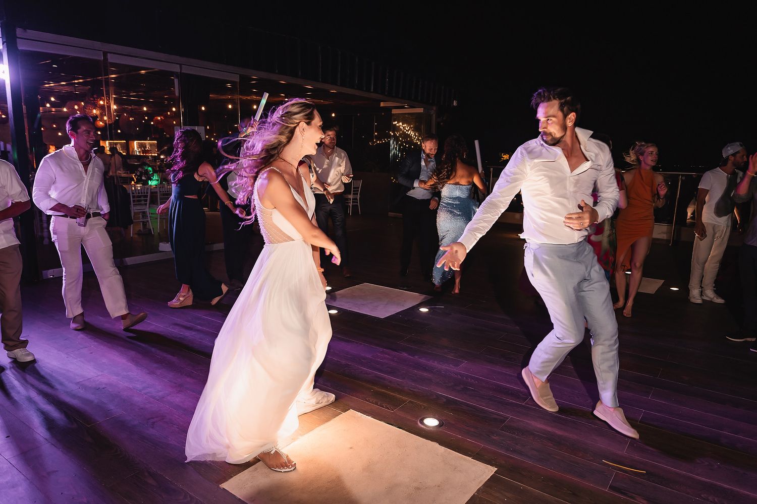 Wedding party dances under purple lighting on dark dance floor during reception.