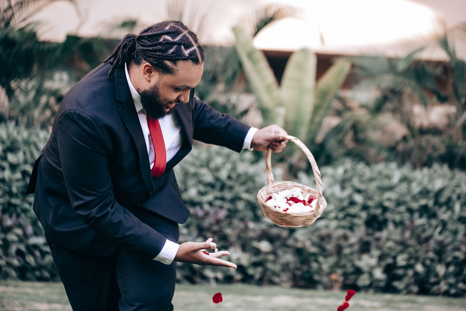 A person in a dark suit and red tie holds a woven basket while standing in a garden setting.
