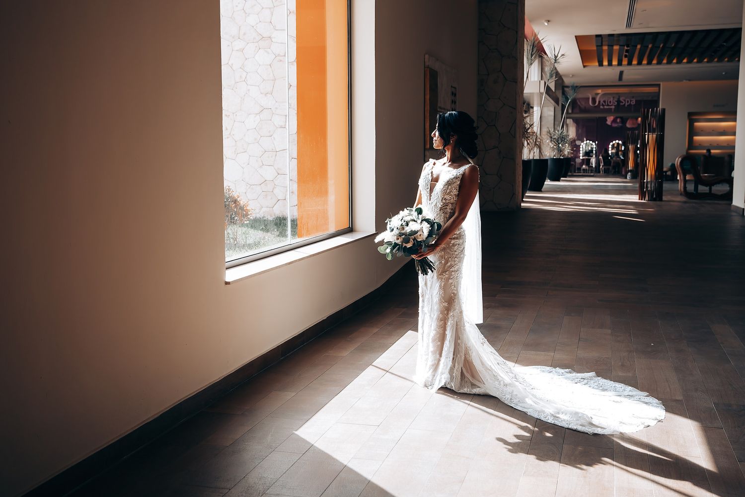 A bride in a white wedding dress walks through a sunlit hallway with dramatic shadows and architectural details.