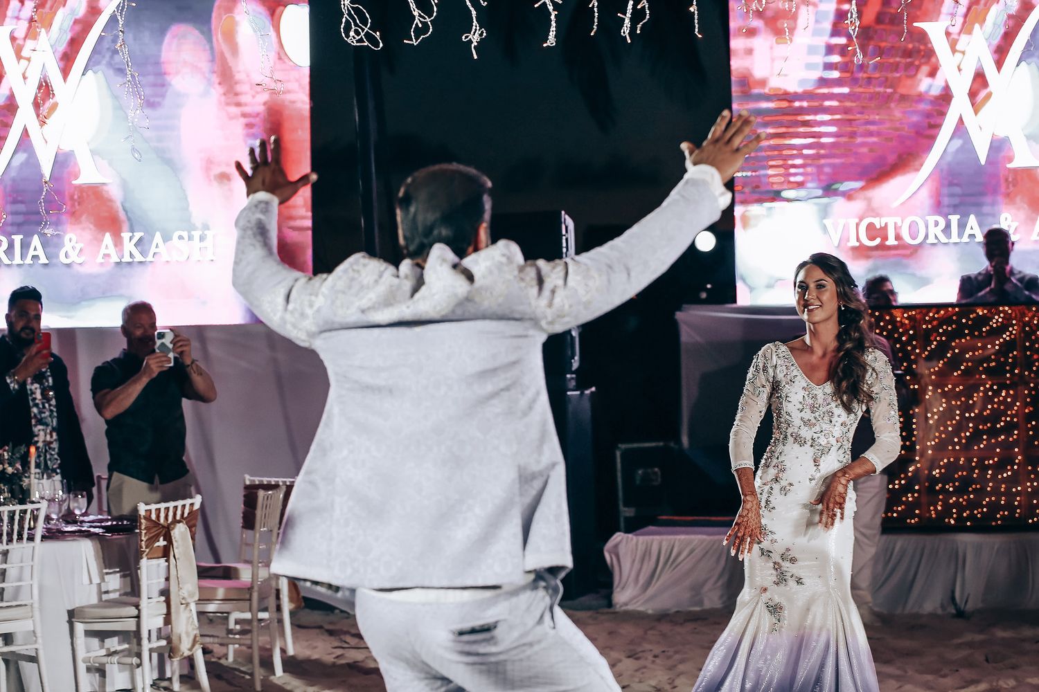 Wedding guests dance and celebrate on a dimly lit dance floor decorated with pink and white lighting effects.