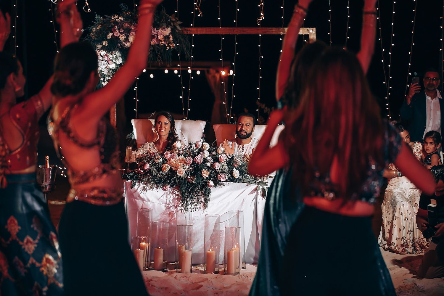 People dancing at a dimly lit wedding reception with red lighting and white chairs in the background.