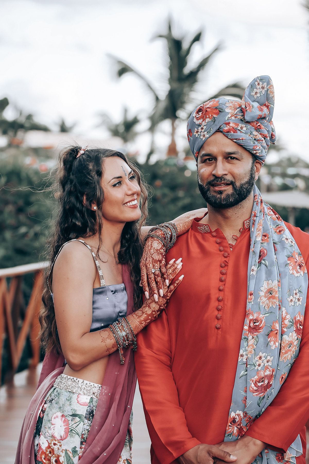A couple in traditional Indian attire share romantic moments outdoors, with the woman wearing floral prints and man in orange.