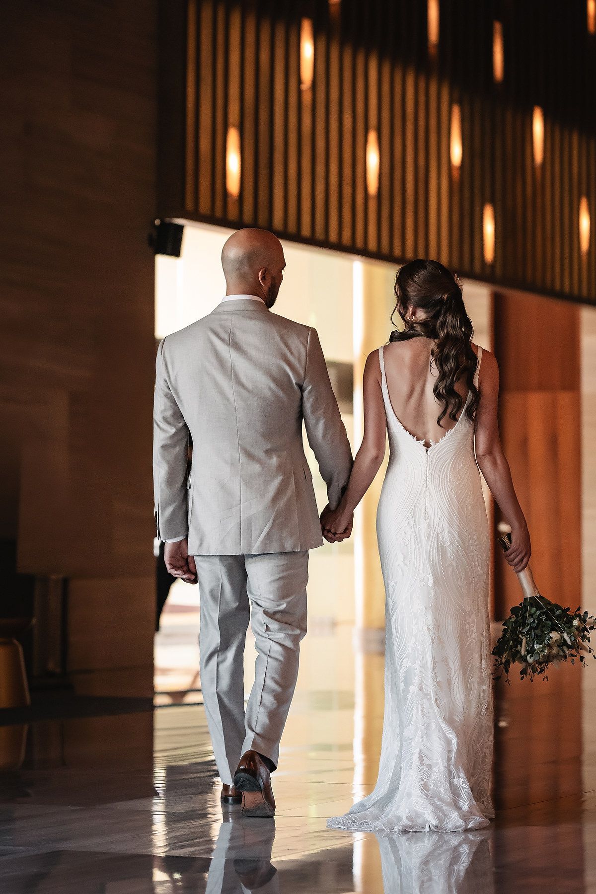 A couple in wedding attire walks through an elegant hallway with warm lighting and wooden paneled walls.