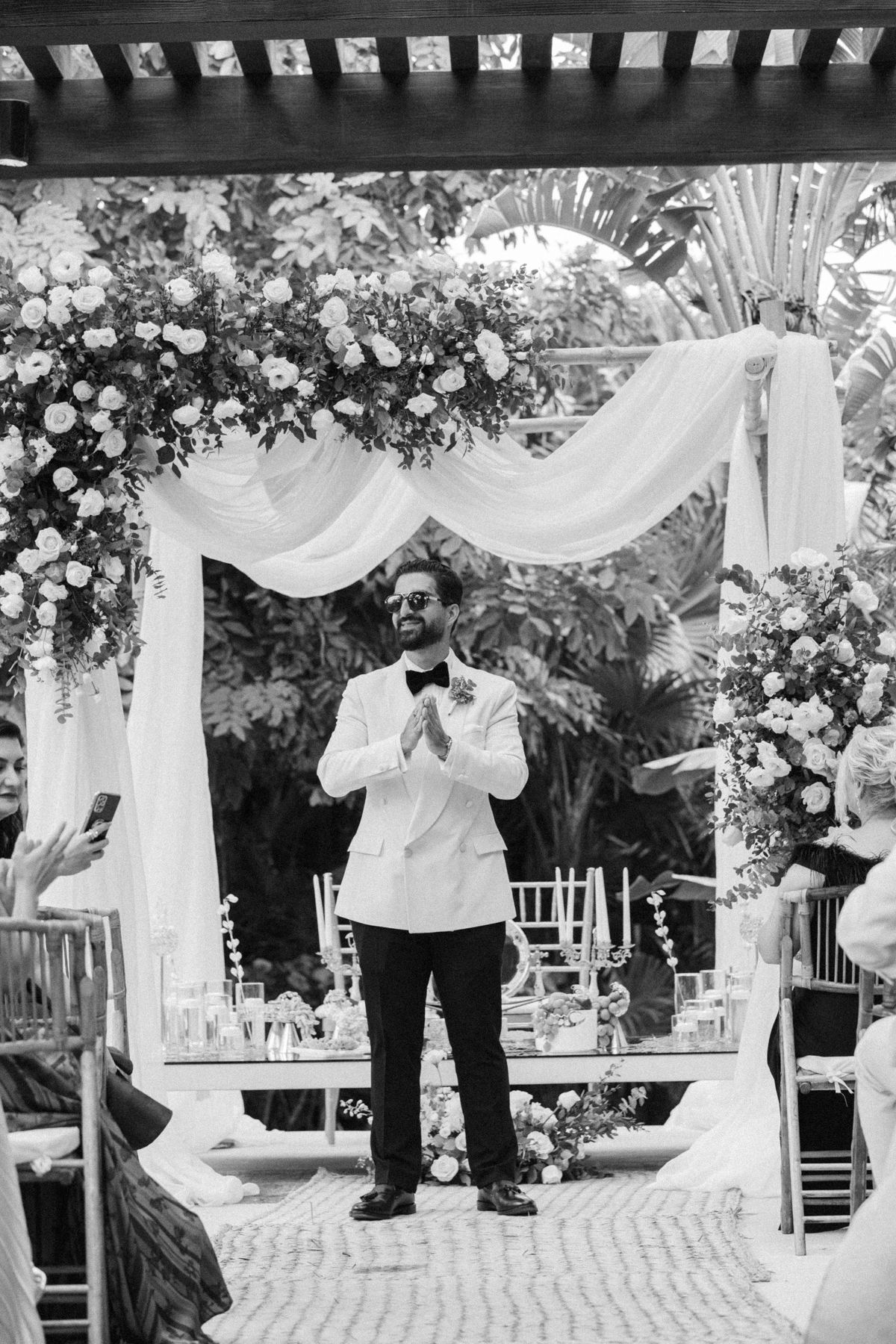 Groom in white tuxedo jacket clapping hands at outdoor wedding ceremony with floral arch and draped fabric backdrop.