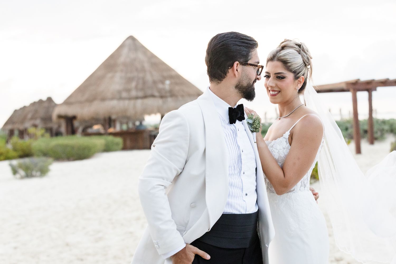 Bride and groom share a romantic moment on a sandy beach with a thatched-roof pavilion backdrop at their destination wedding.