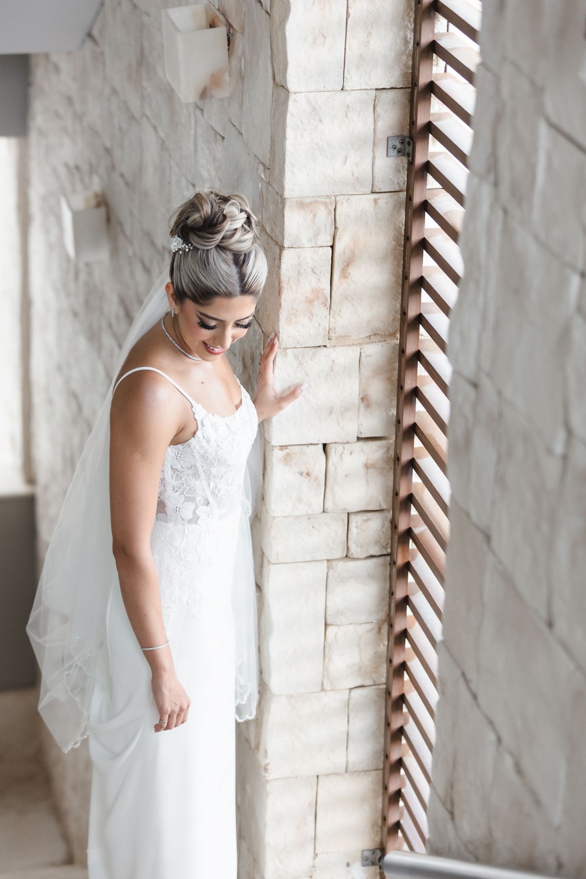 Bride in white lace gown and veil poses gracefully by a stone wall near a wooden window shutter.