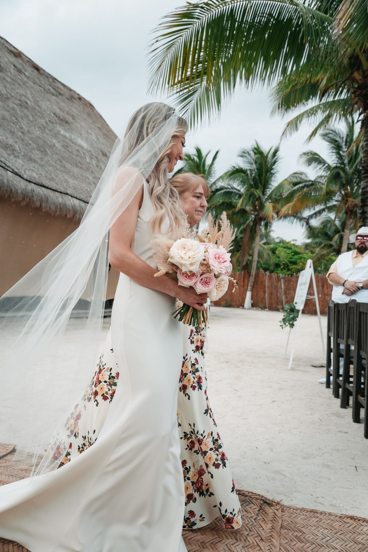 Bride in white gown and veil shares an emotional hug with her mother in a floral dress on the beach.