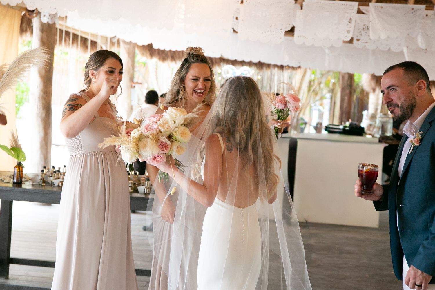 Bridesmaids in champagne gowns celebrate with bride in white wedding dress under thatched reception venue.