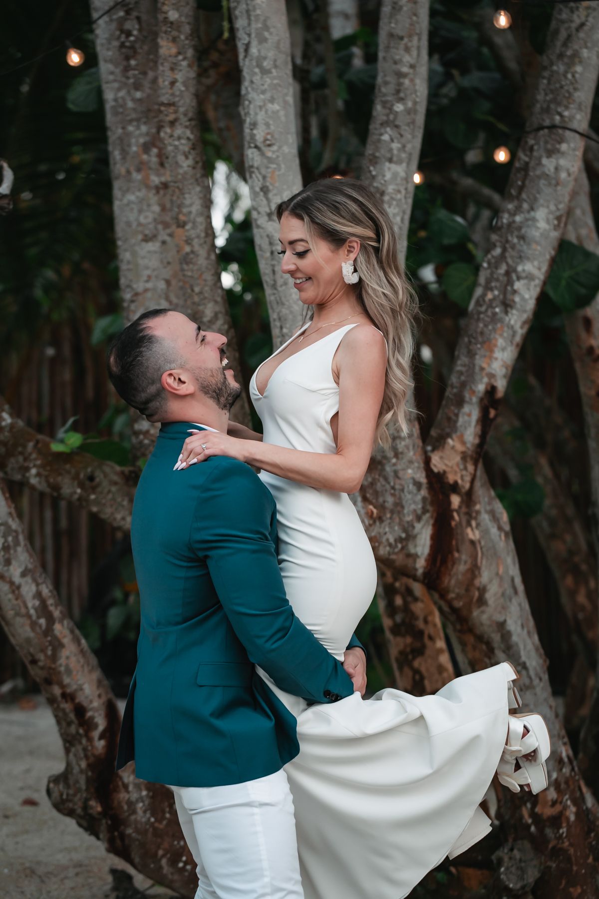 Groom lifts bride in white fitted gown among tropical trees with string lights at romantic outdoor wedding.