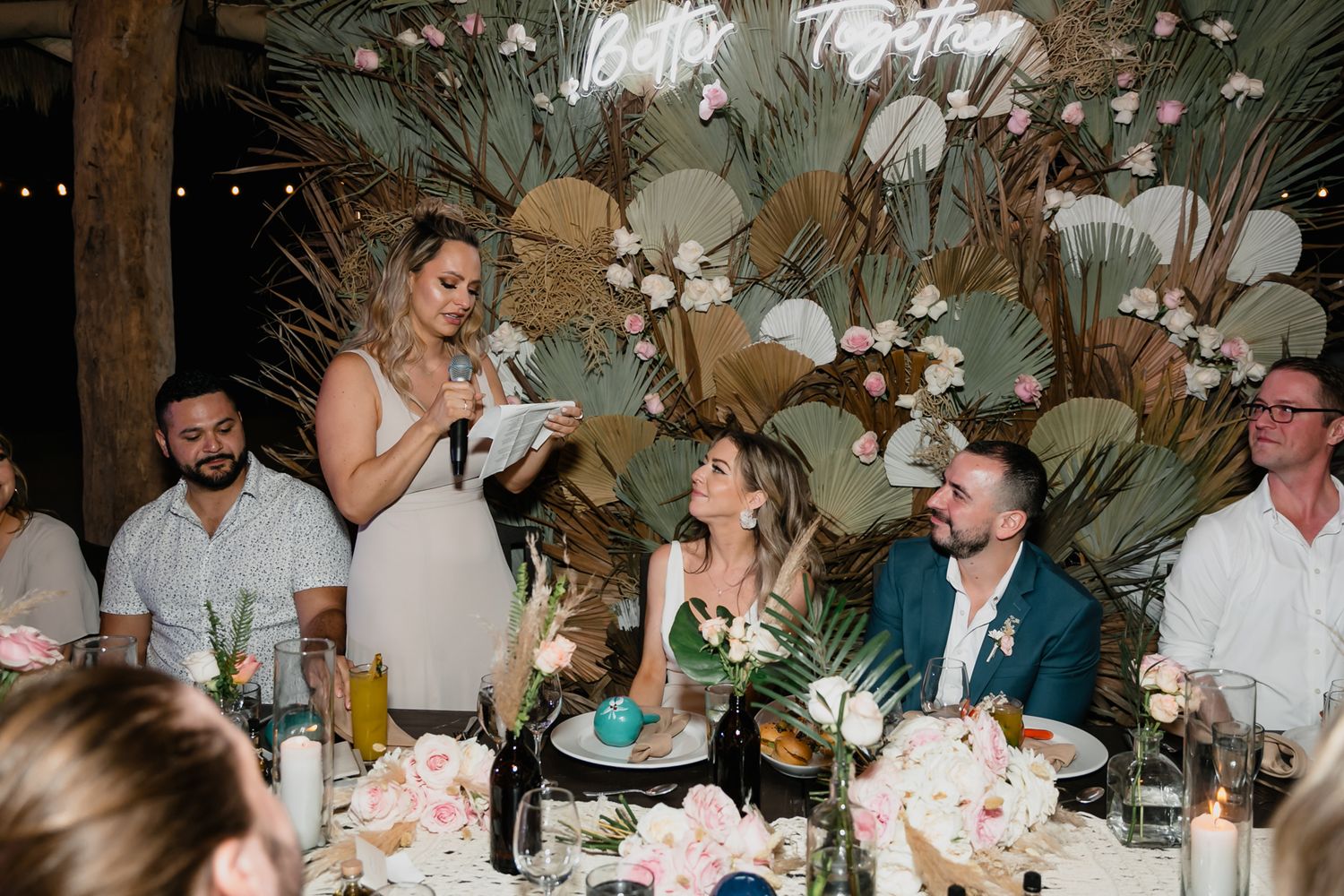 Bridesmaid gives heartfelt speech at tropical wedding reception as bride and groom listen at floral decorated head table.