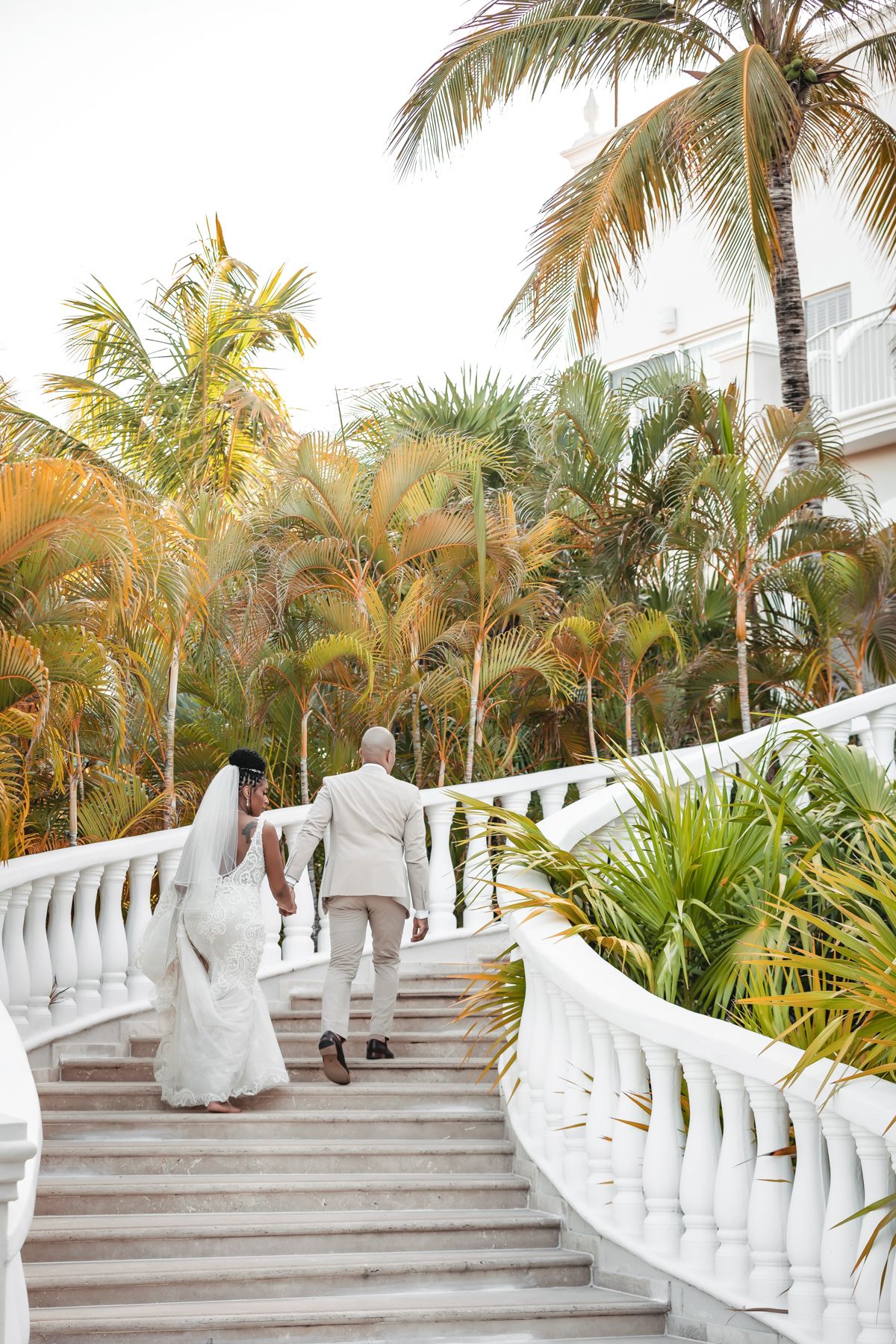 Bride and groom walking up white marble staircase surrounded by tropical palm trees at a destination wedding venue.