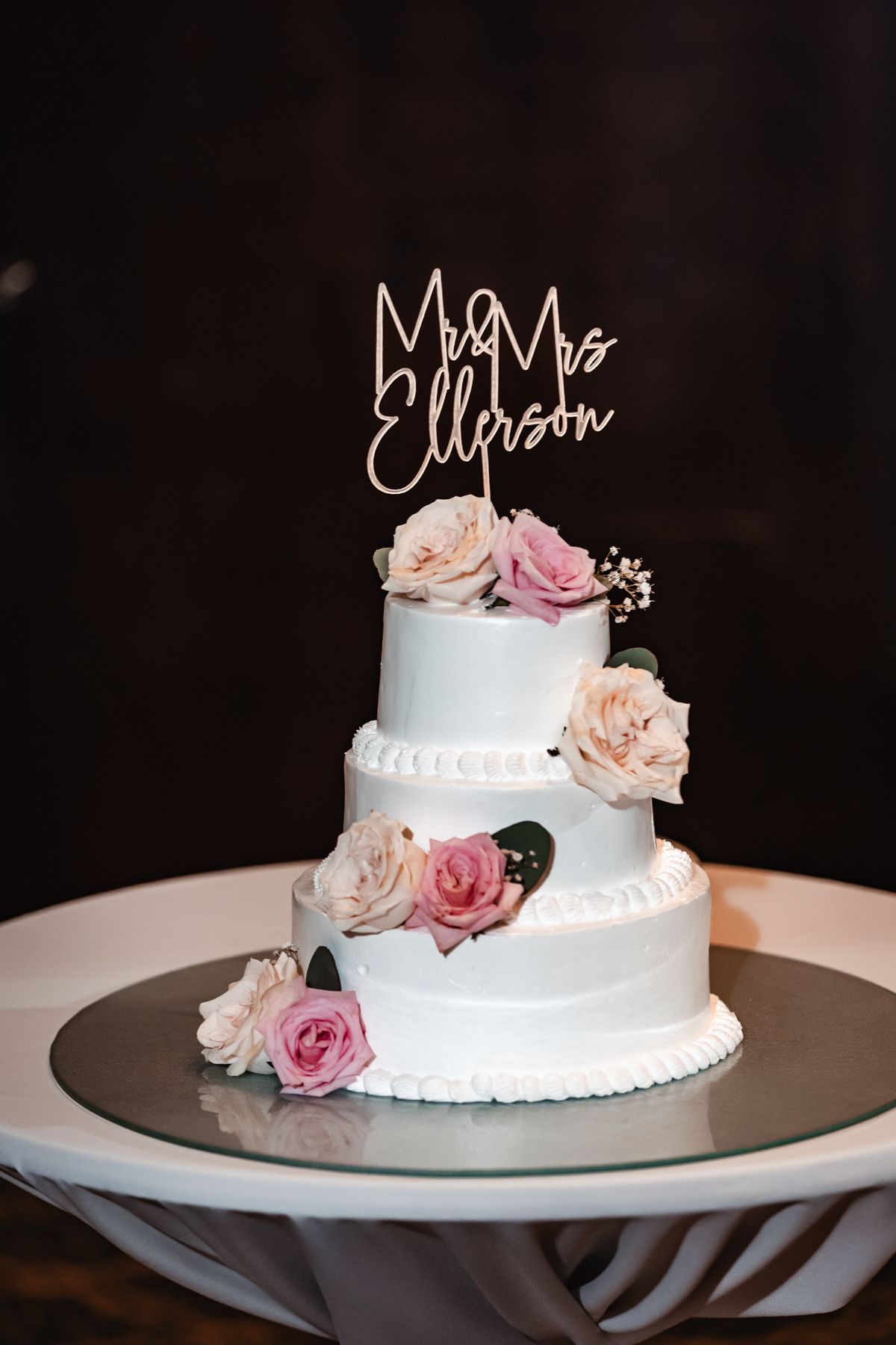 Three-tier white wedding cake with pink roses and Mr & Mrs Ellerson gold cake topper on a elegant cake stand.