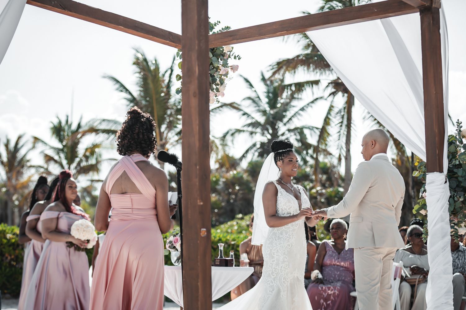 Bride and groom exchange vows under a wooden arch with bridesmaids in pink dresses at an outdoor beach wedding.